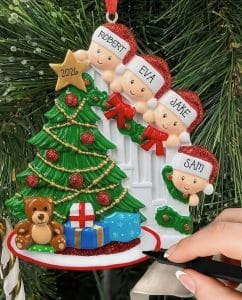 Decorative Christmas ornament: four children’s heads in Santa hats around a festive tree with gifts and a teddy bear, held by a hand with a pen nearby.