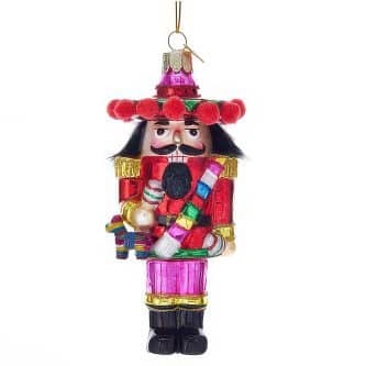 Colorful nutcracker ornament with a red coat, gold trim, and a pink hat adorned with red pom-poms, hanging by a string.